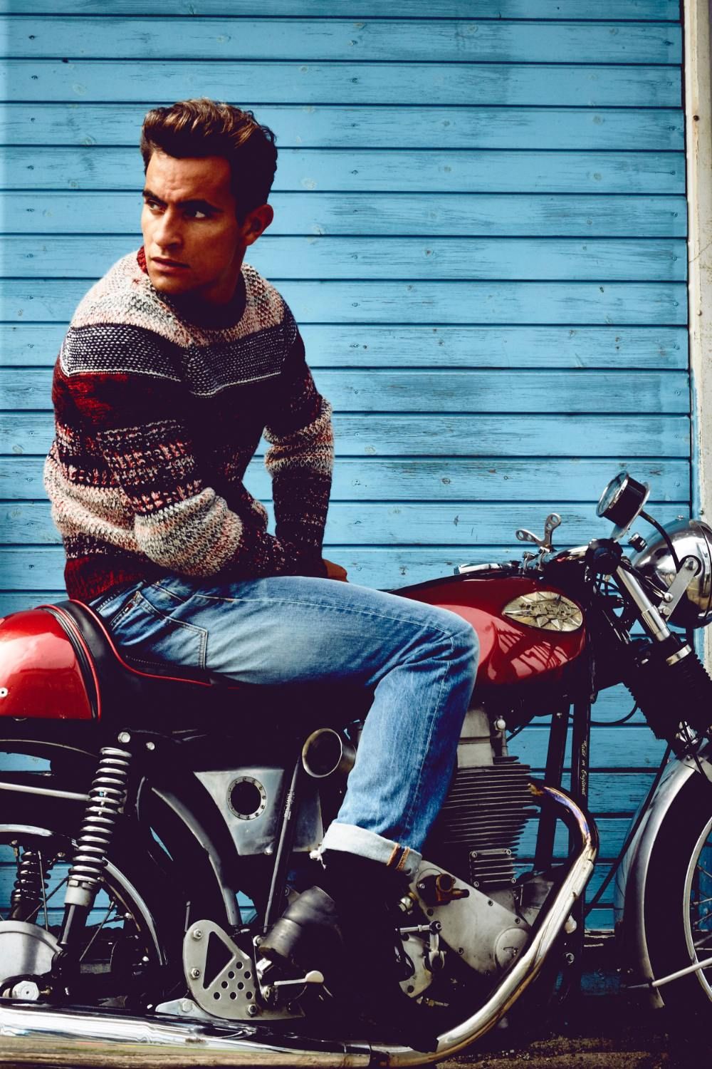 Jonathan Harboe sits on a red motorcycle against a blue wooden backdrop, exuding a contemplative mood.