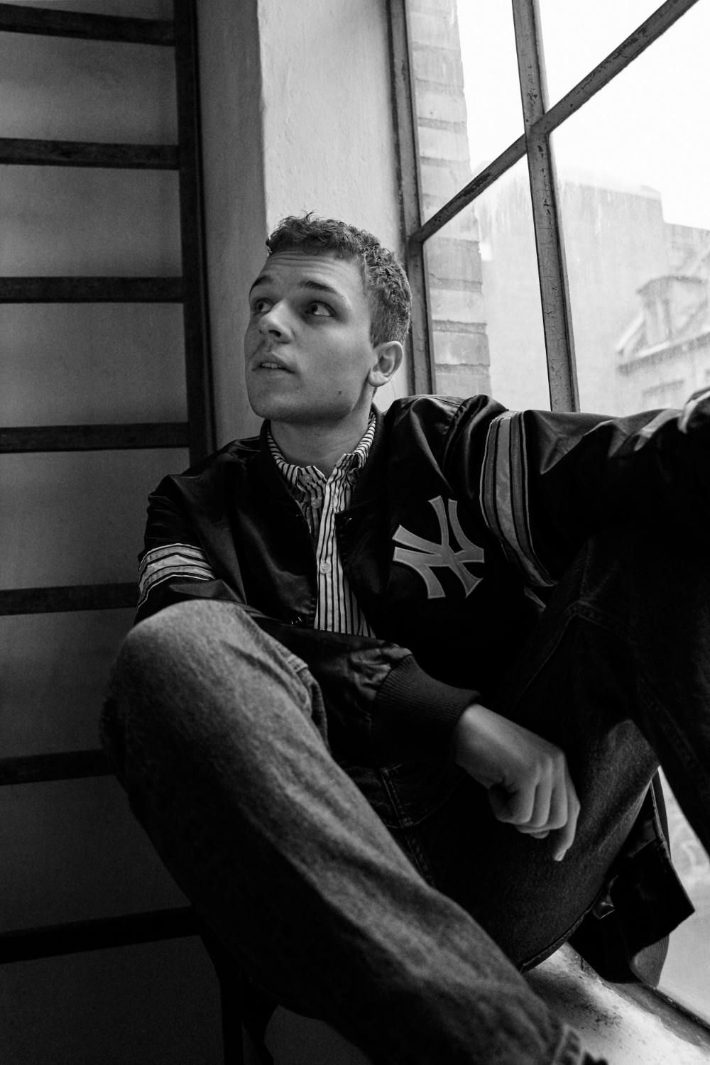 Rasmus Brohave seated by a window, wearing a jacket and looking thoughtfully upward.