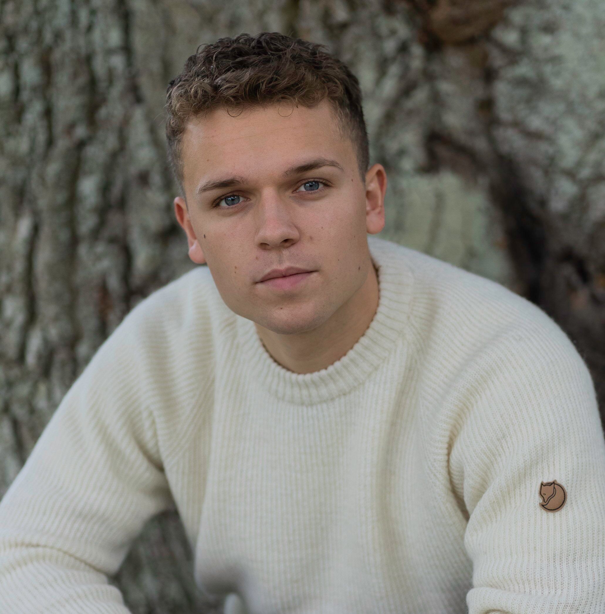 Rasmus Brohave poses thoughtfully against a textured tree trunk, wearing a cream sweater.