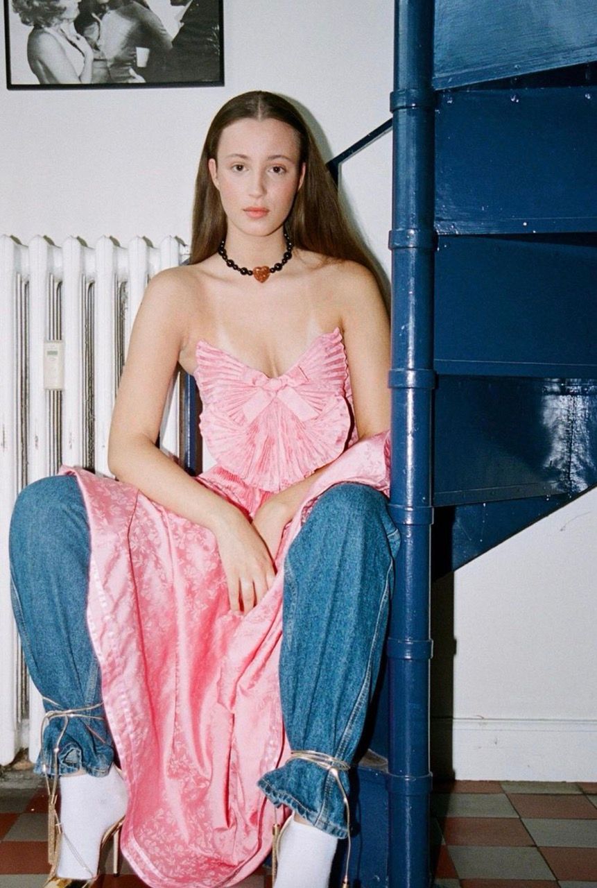 Josephine Højbjerg seated on stairs, wearing a pink dress and denim, with a neutral expression.