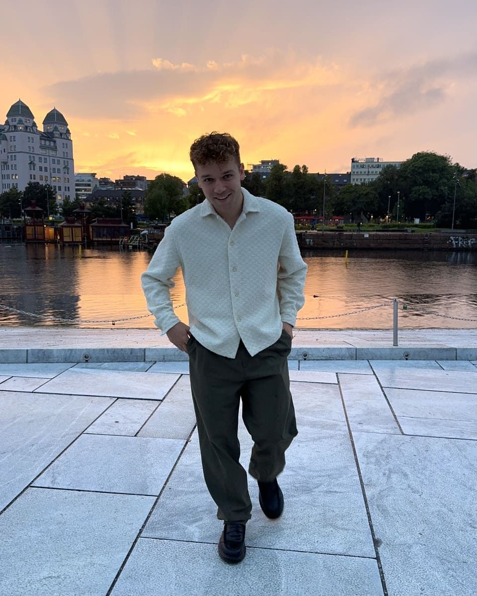 Rasmus Brohave poses by a waterfront at sunset, wearing a light shirt and dark pants.